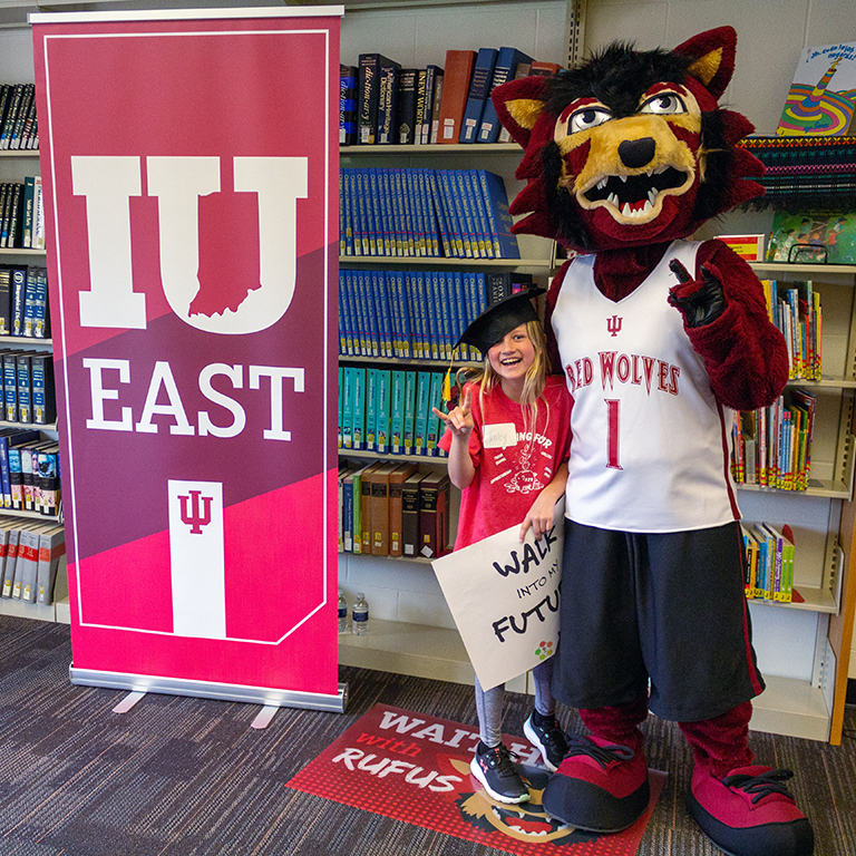Rufus the Red Wolf posing with a third grader in school library beside an IU East banner