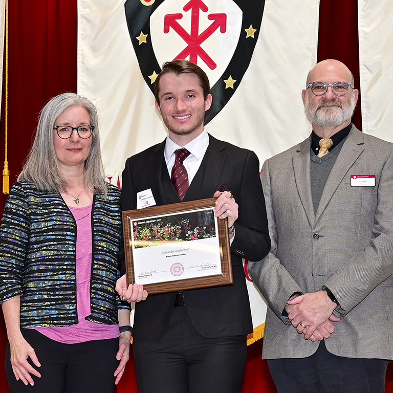 Alex Austerman holding his framed certificate between IU East administration in front of a banner