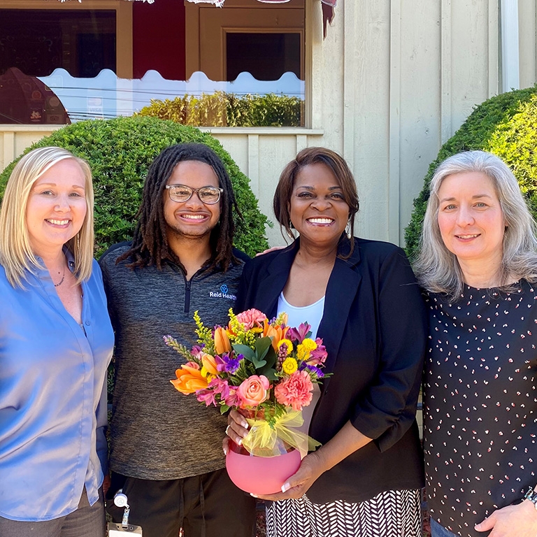 Four people standing together, smiling. Sharrie Harlin on the center-right is holding a bouquet of assorted, colorful flowers.