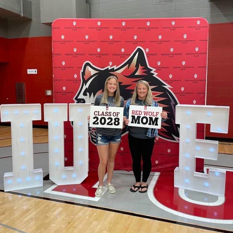 Mother and daughter standing in front of crimson backdrop and illuminated IU letters, holding signs that read Class of 2028 and Red Wolf Mom