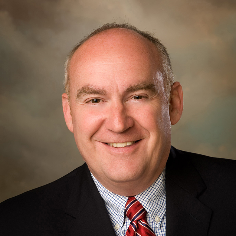 Tim Scales smiling in front of a warm grey background.