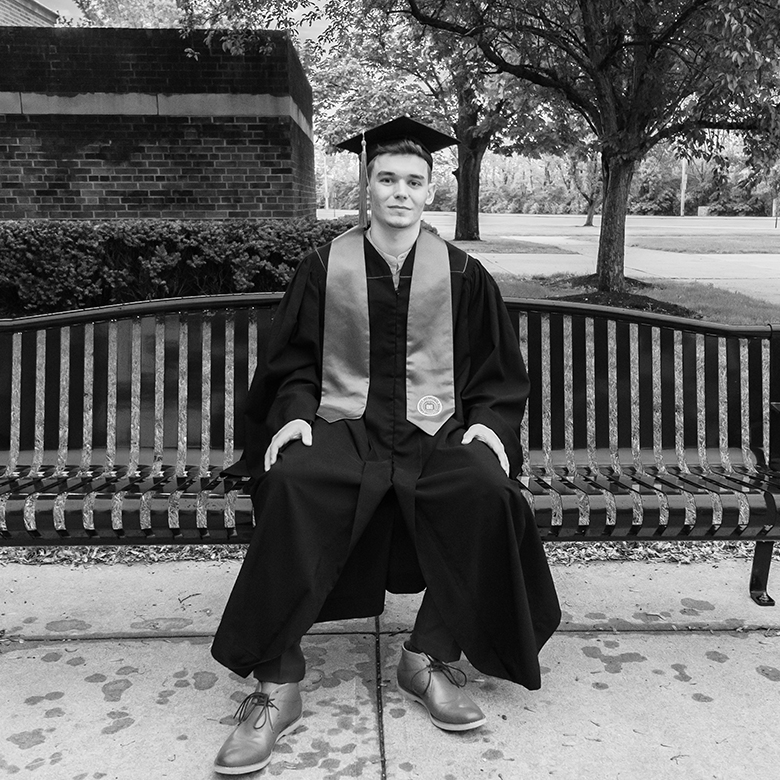 Hayden Jarvis, dressed in a graduation cap, gown, and stole, sitting on a bench.
