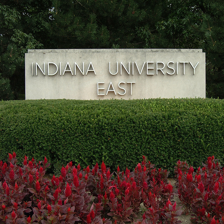 A stone sign depicting the name of the Indiana University East campus, set behind a landscaped shrub and red flowers