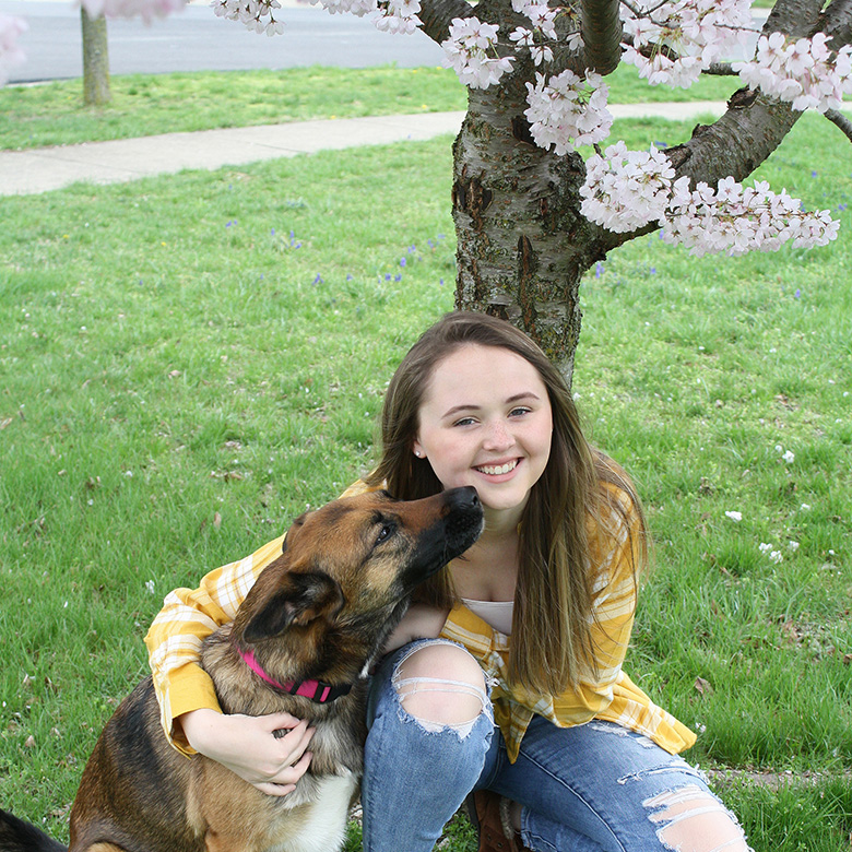 Jillian Splawn, smiling and kneeling under a blossoming tree with an arm around a dog.