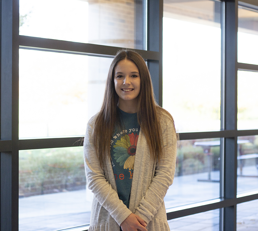 Emma Frye standing in front of windows, smiling with her arms down and hands clasped in front of her