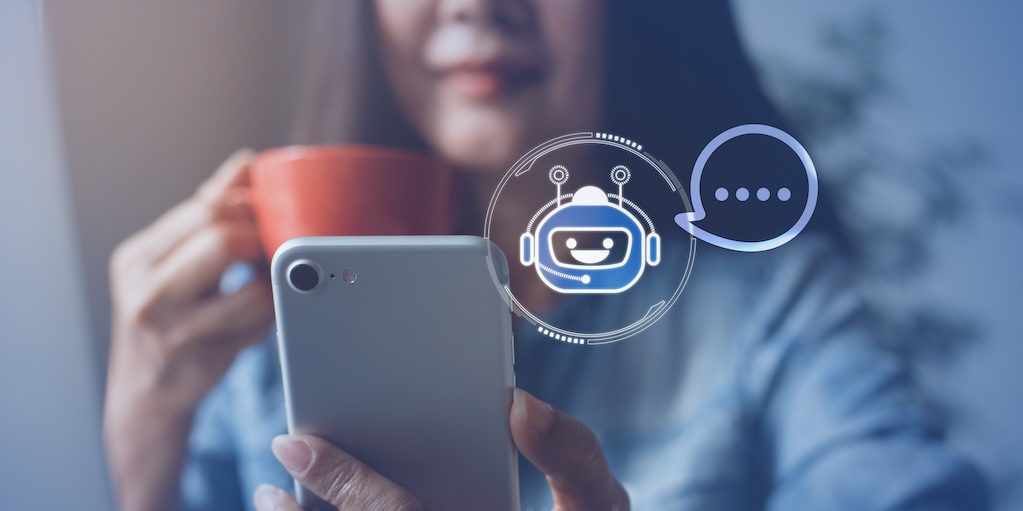 Woman holding tea cup and looking at smart phone in her hand. Small icon of a chatbot with a speech bubble is shown near the phone.