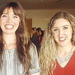 Two women smiling at an award event. The woman on the right, wearing a red dress and statement necklace, holds a framed certificate recog...