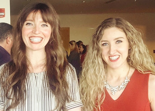 Two women smiling at an award event. The woman on the right, wearing a red dress and statement necklace, holds a framed certificate recog...