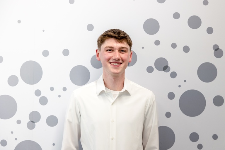 A young man wearing a white button-down shirt stands smiling in front of a white wall decorated with various sizes of gray circles. He ha...