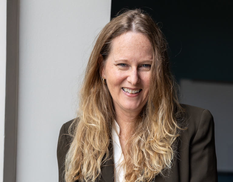 Smiling woman with long, wavy blonde hair wearing a dark blazer and white blouse, standing indoors near a light-colored wall.