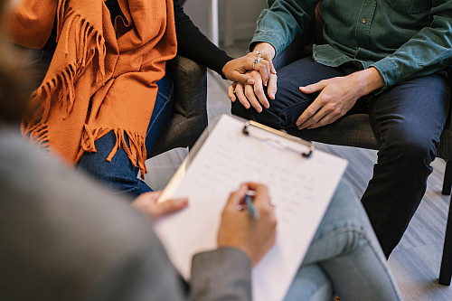 Detailed crop of hands of anonymous husband and wife sharing marriage problems and holding hands while therapist taking notes in notepad ...