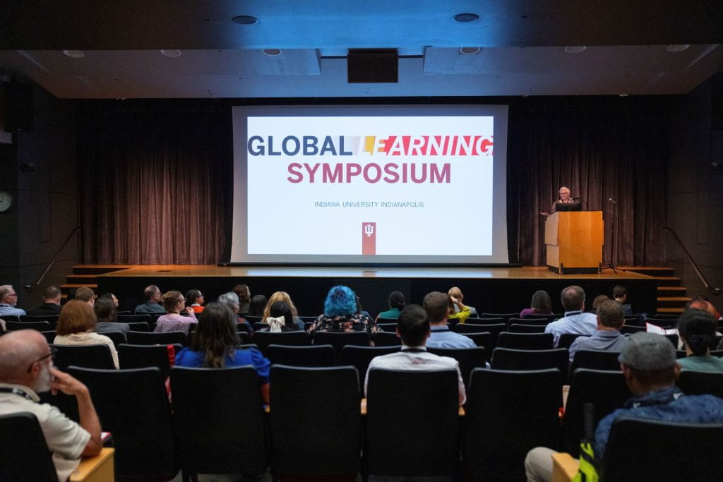 The view of a stage from the audience. A screen reading Global Learning Symposium is visible over the heads of audience members. Associat...