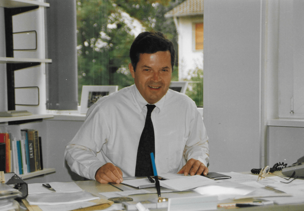 A portrait of a young Wolfgang Dexheimer, a man with brown hair wearing a white shirt and black tie.