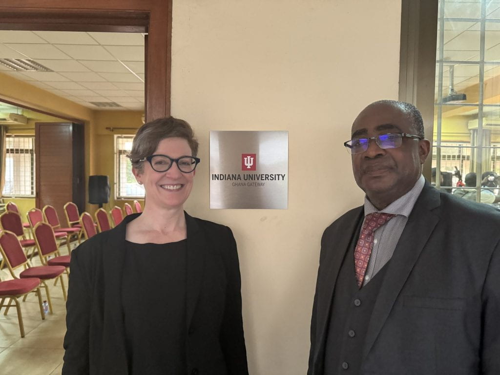 Two people smile at the camera. Between them a plaque on the wall reads Indiana University Ghana Gateway