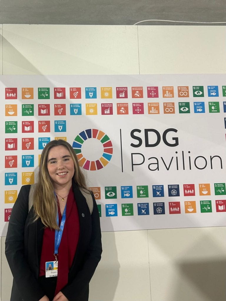 Gie Wilson, a student with long hair wearing a blouse, backpack, and blazer, stands in front of the SDG Pavillion. She wears a COP29 badg...