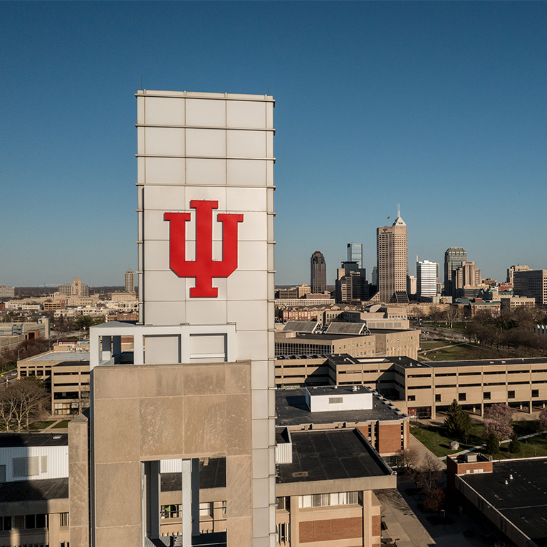 Photo of the top of the Bell Tower of the Campus Center that features a crimson IU trident at the top. The Indianapolis skyline is in the...