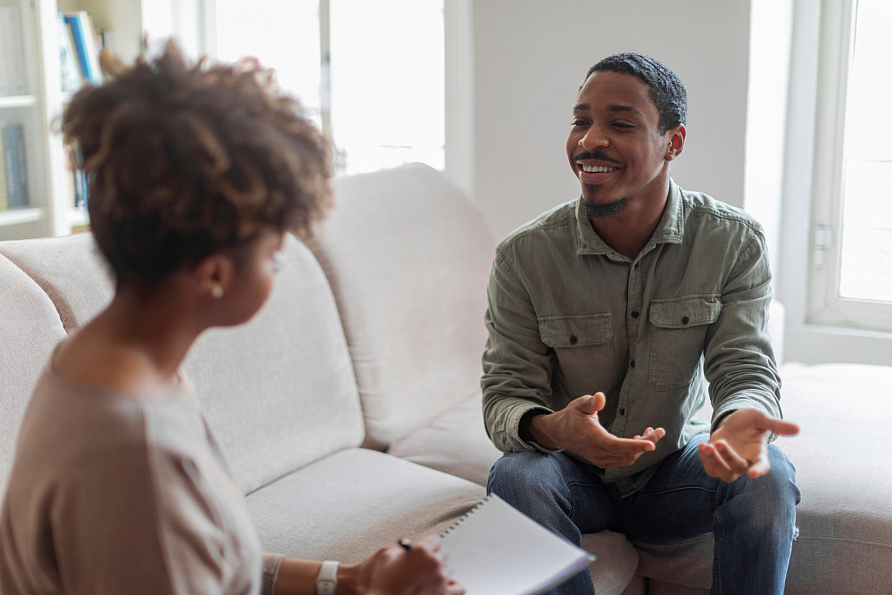 Cheerful positive handsome young african american guy in casual outfit sitting on couch at cozy counselor office, having conversation wit...