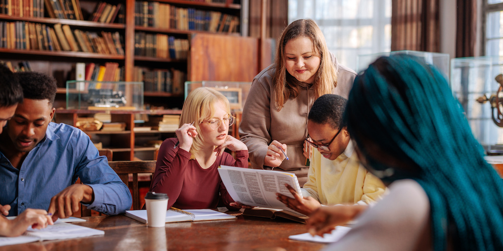 Students seated and talking at a table in library, bookshelves behind.